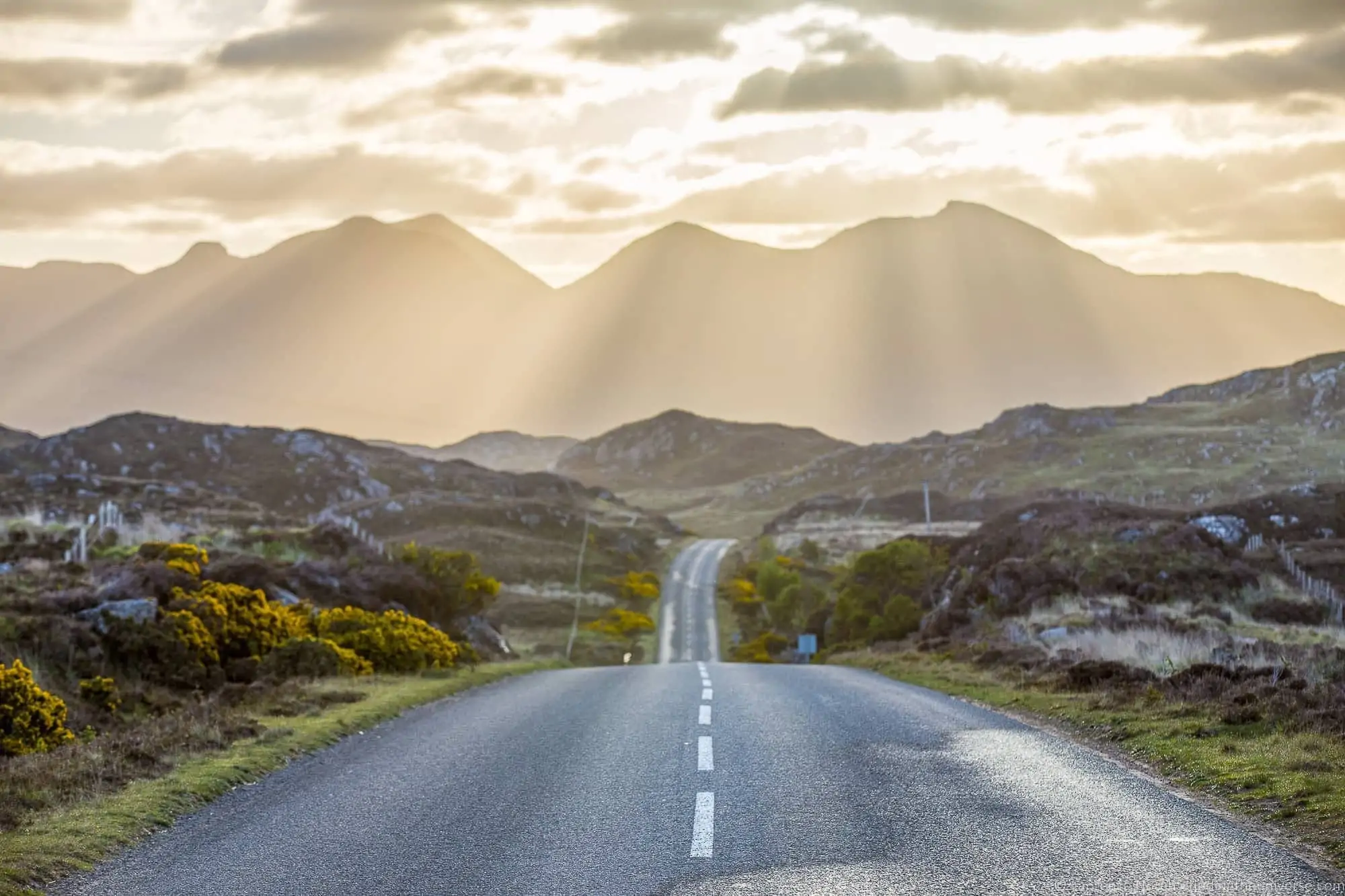 Sunbeams breaking over the mountains above an empty road on Scotland's North Coast 500