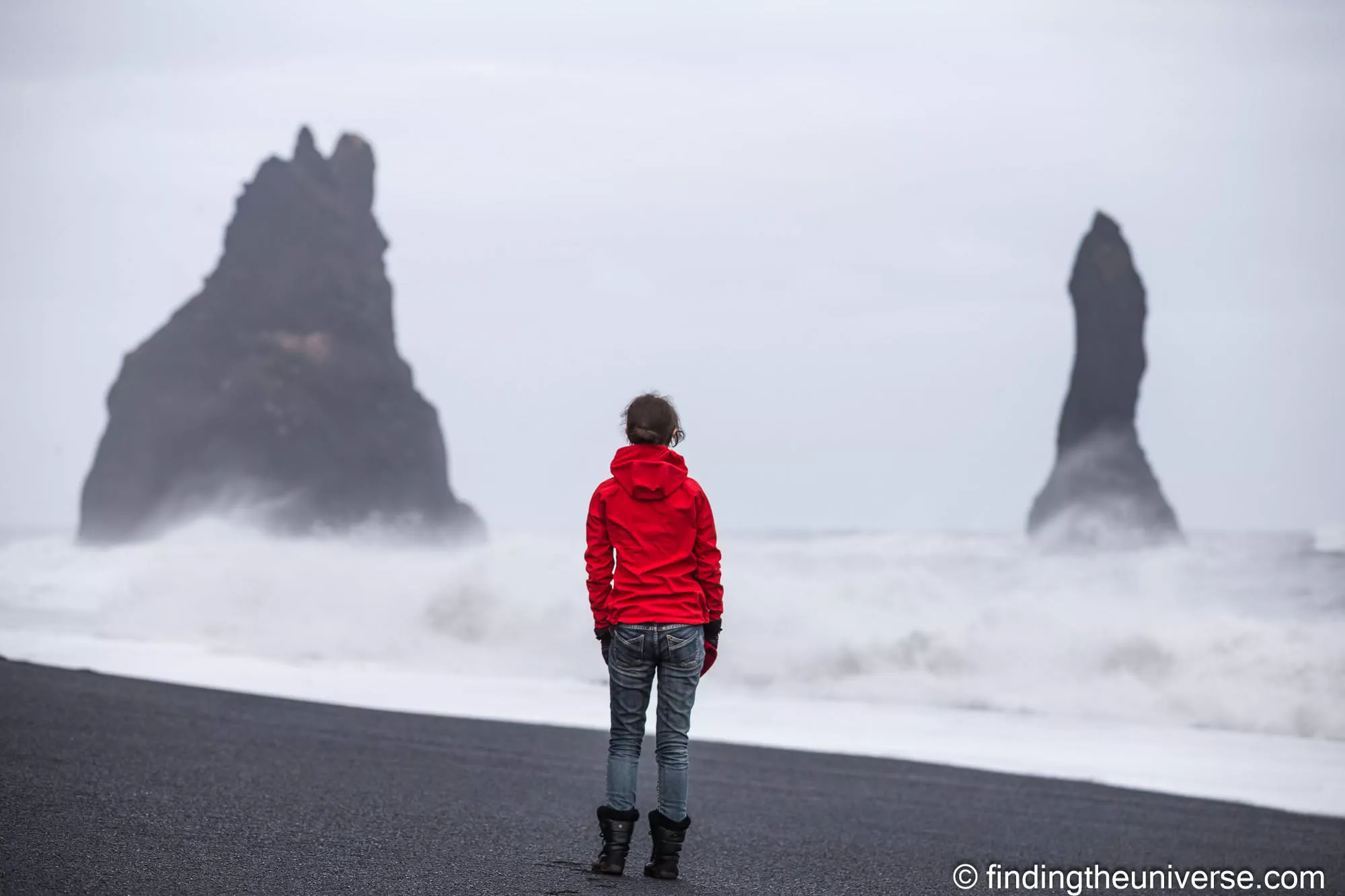 Person in a red jacket looking out at the Reynisdrangar sea stacks from Reynisfjara black sand beach
